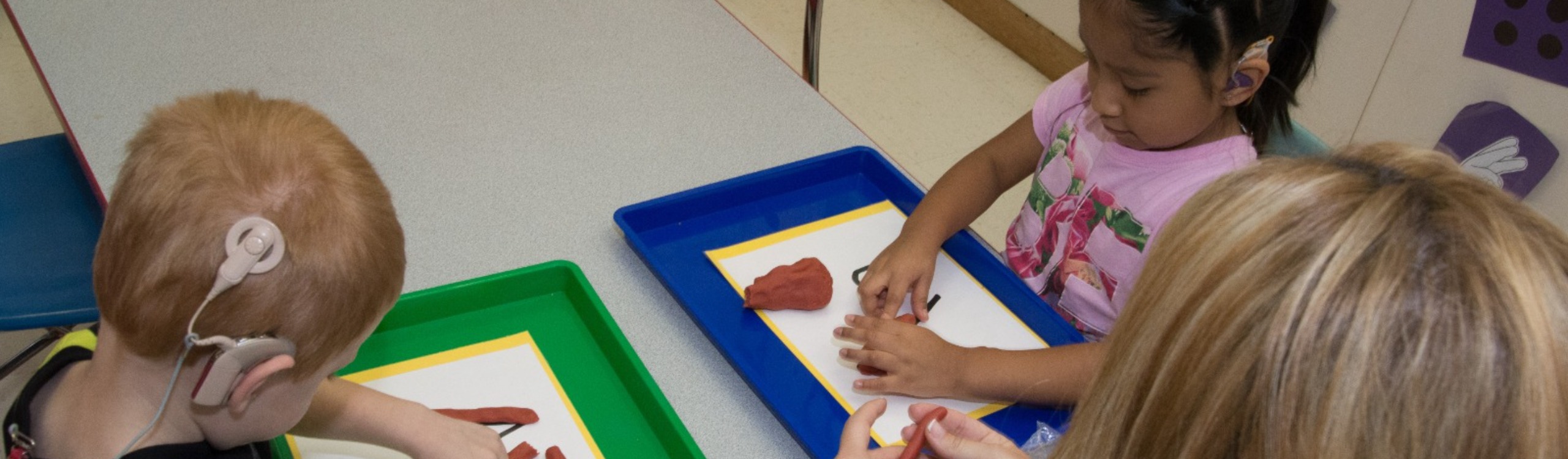 One boy and one girl seated facing each other at a table. Both are making letters using clay. A teacher is seated at end of table assisting.