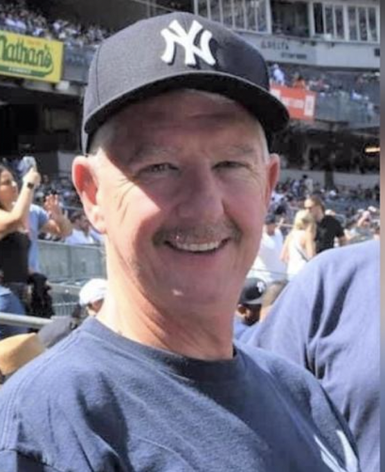 Greg Page smiling, wearing a Yankees hat and Yankees t-shirt, with Yankee Stadium stands in background.