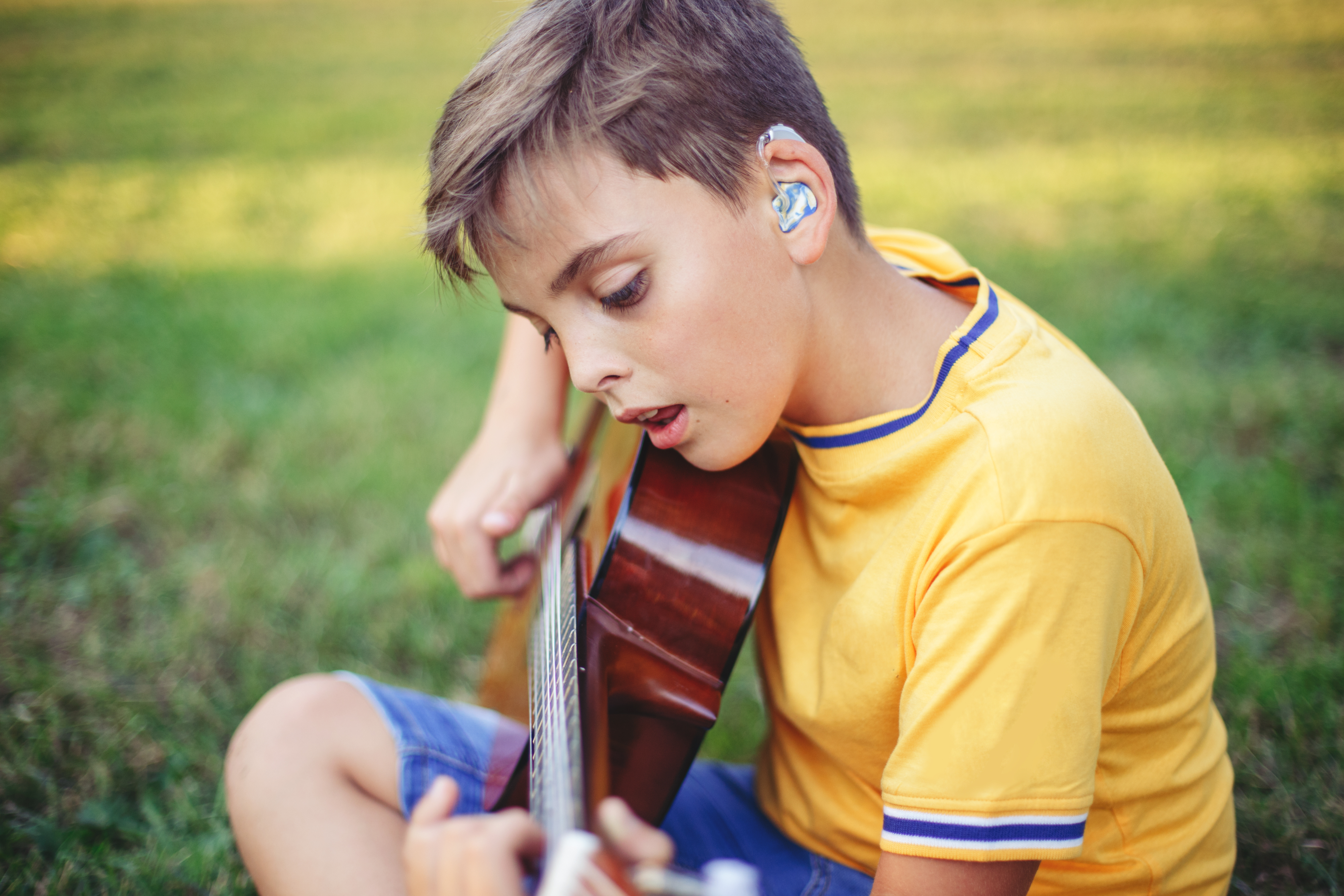 Student with hearing aid playing the guitar while sitting in the grass