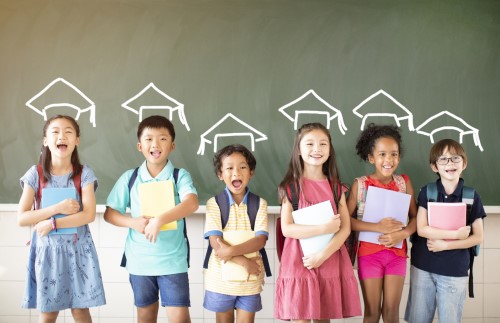 Elementary students holding books in front of chalkboard with graduation caps drawn above their heads