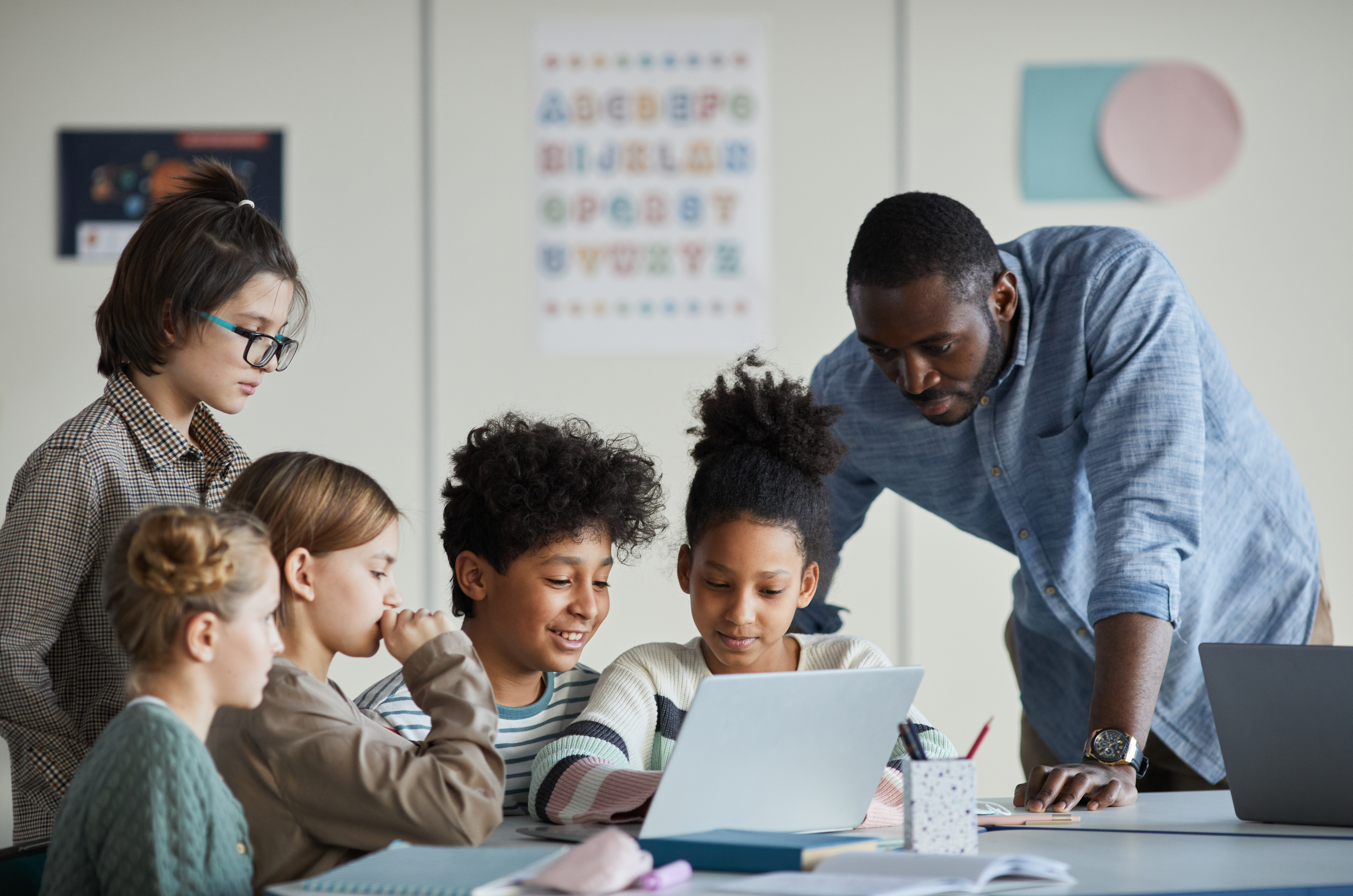 Elementary students sitting at table looking at laptop with teacher