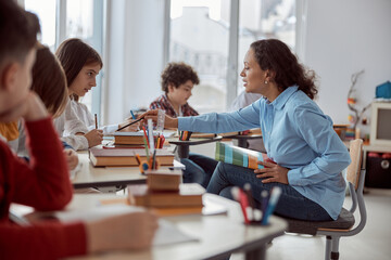 Young teacher helps to read her student. Elementary school kids sitting on desks and reading books in classroom