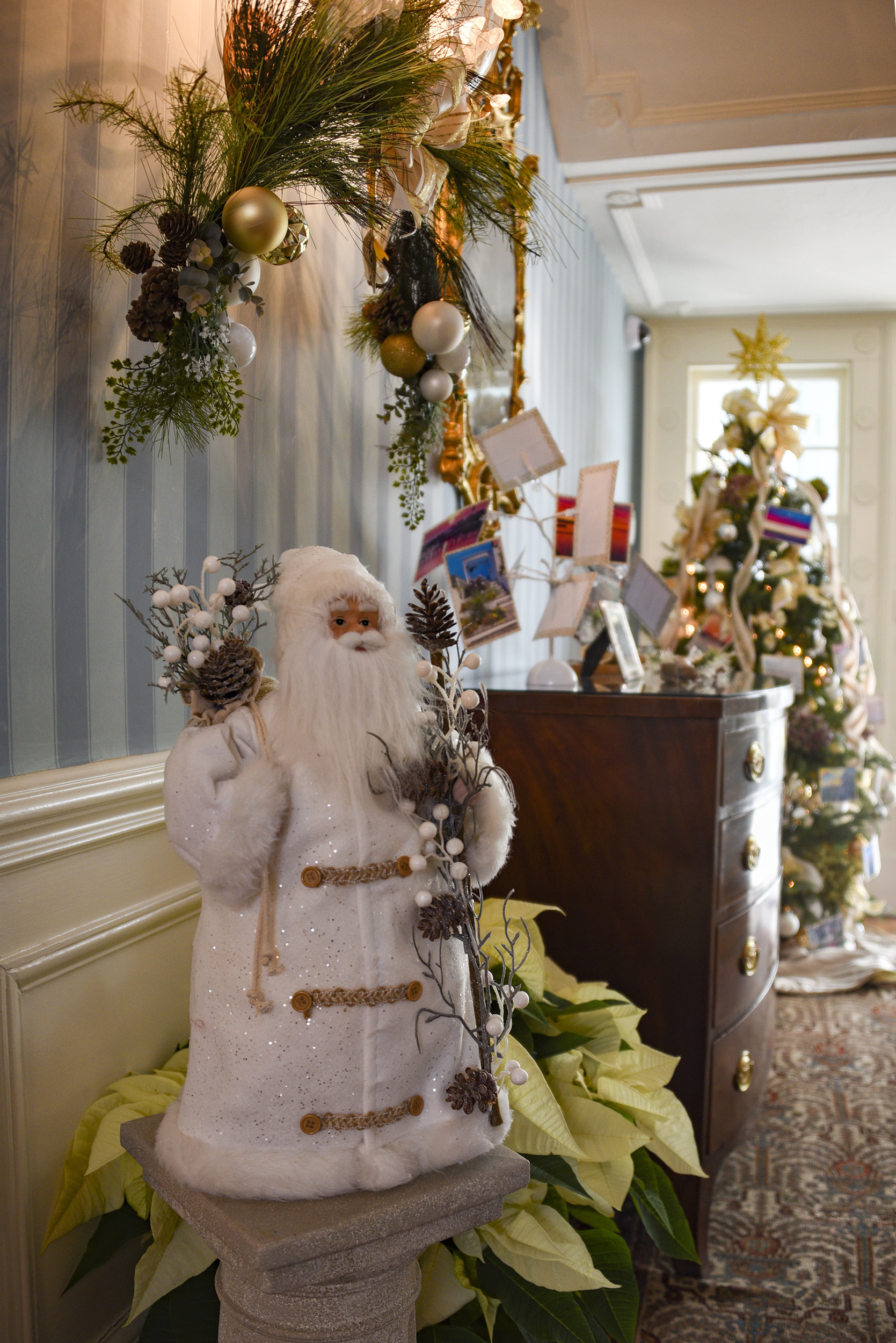 The Foyer, by Keyport Garden Club, features numerous elegant decorations including a white-robed Santa Claus figure and a large wall spray of greenery with gold and cream colored balls and pinecones.