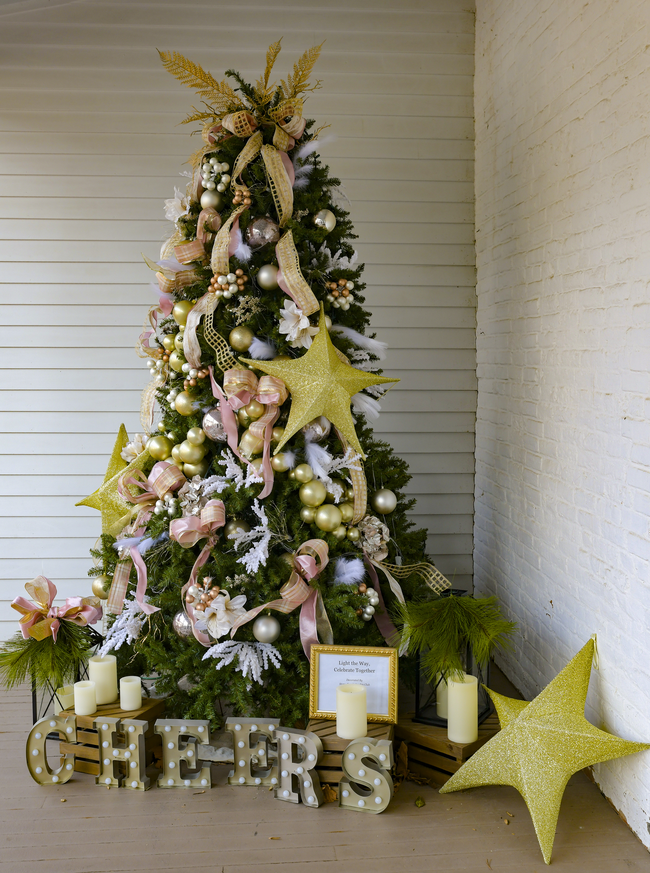 Stony Brook Garden Clubs “Light the Way” Christmas tree on Morvens Back Porch features gold and cream balls and pink ribbons and bows. Displayed are gold stars, electric candles and large letters at the base of the tree spelling out the word "cheer