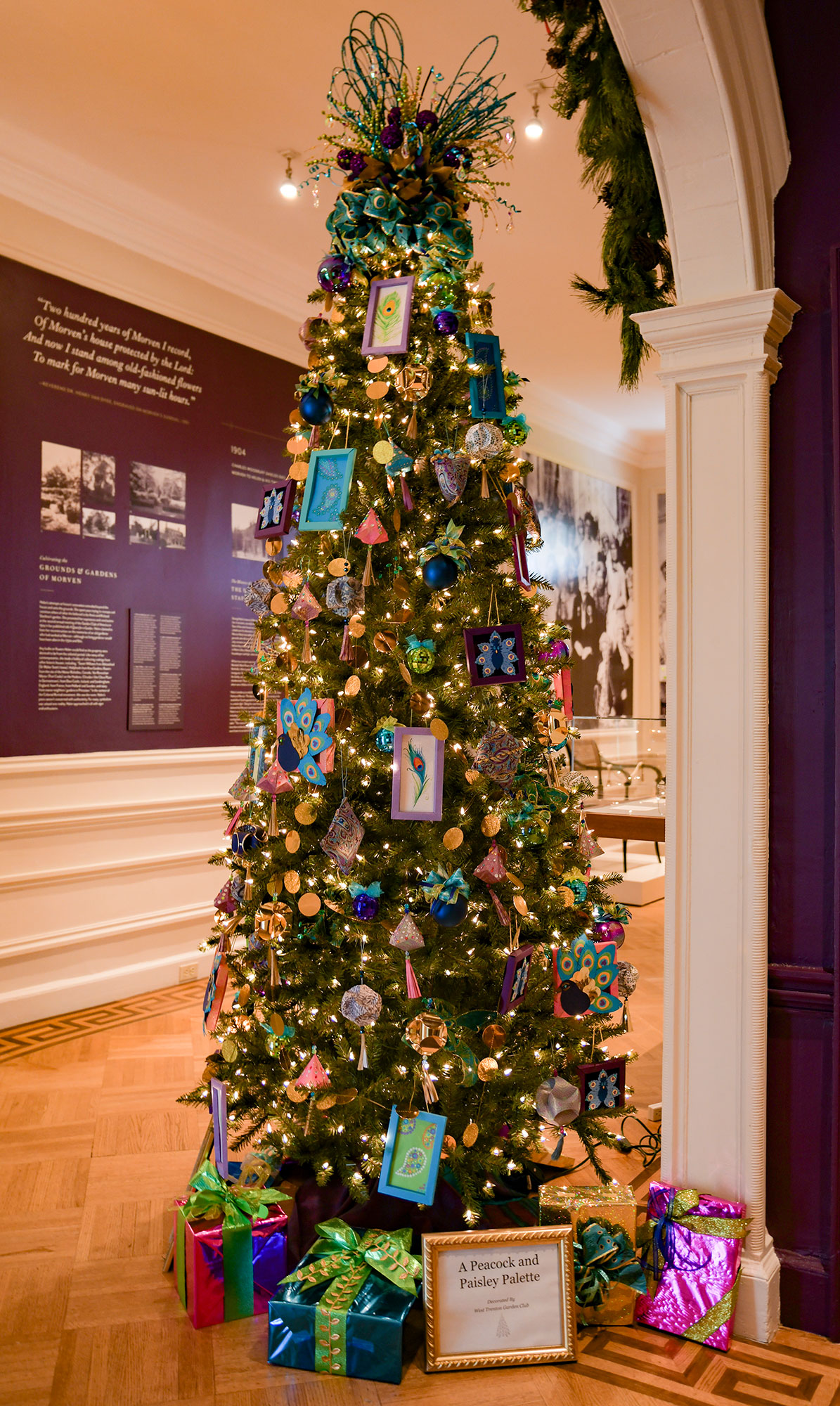 A “Peacocks and Paisley Palette Christmas” tree by West Trenton Garden Club is seen in the Foyer with many vibrant colors. On the walls across and behind are historical displays.