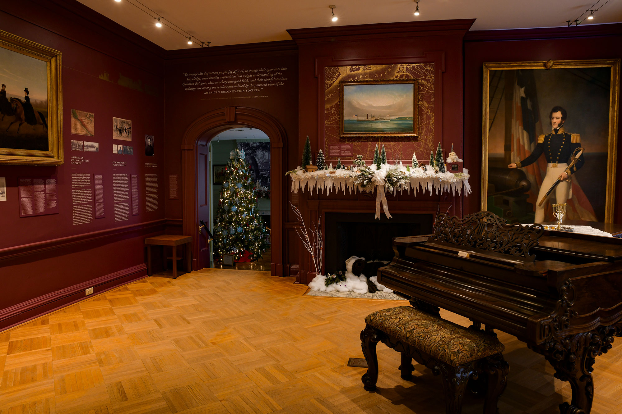 A winter-themed mantelpiece by Neshanic Garden Club is shown in the music room. Plum colored walls, historic paintings and a grand piano are prominent. In the background entry to another room where a Christmas tree is lit.