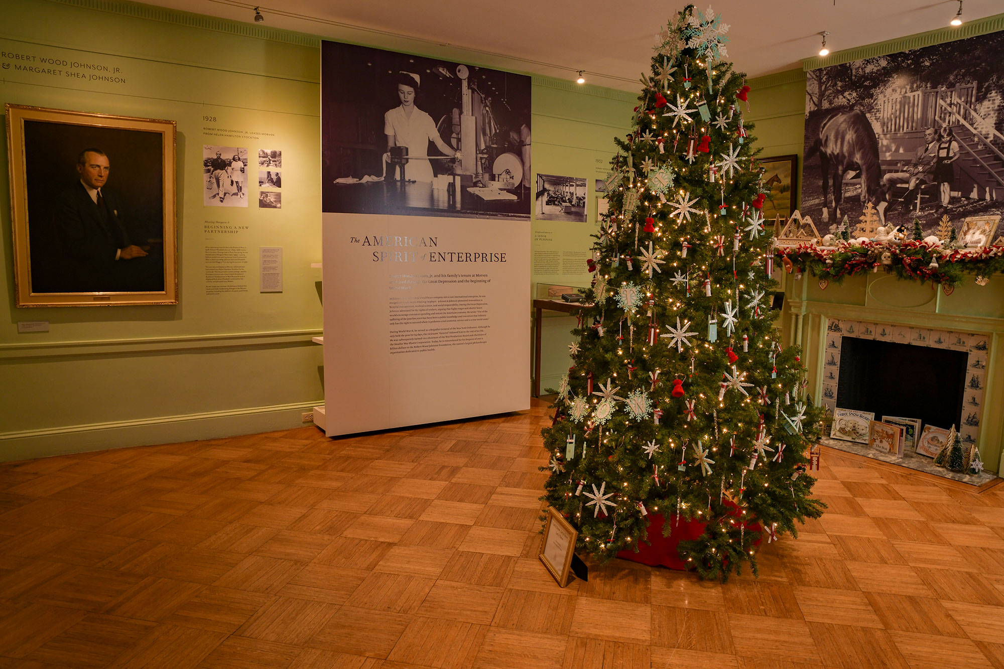 A Christmas tree in a light green room with a fireplace, biographical displays and decorated mantelpiece. The Garden Club of Princeton tree features snowflake ornaments and the mantelpiece by Princeton Public Library features books by Jan Brett.