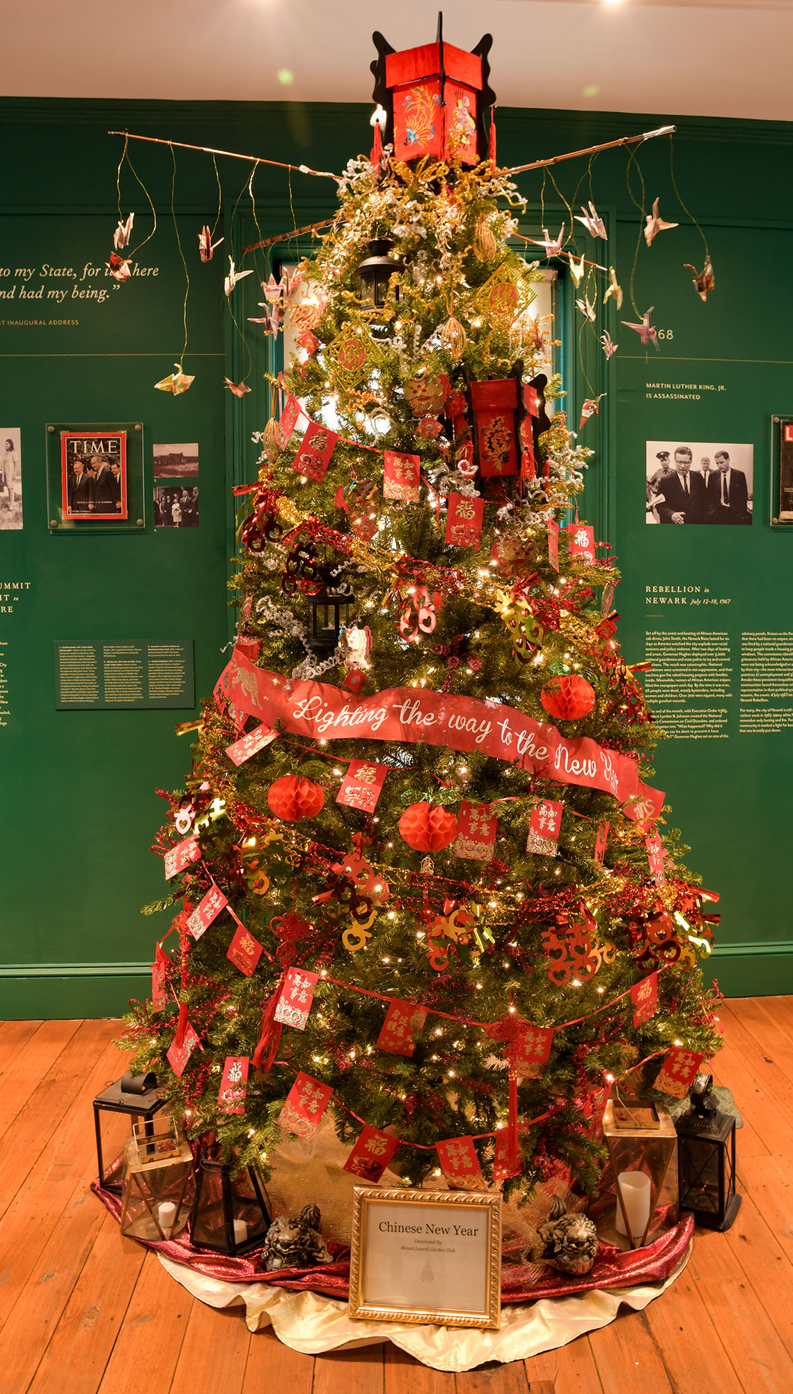 Mount Laurel Garden Club tree called “Chinese New Year” is seen in a room with dark green walls featuring historical information. Bright red garlands wrap around with the center ribbon reading Lighting the Way to the New Year.