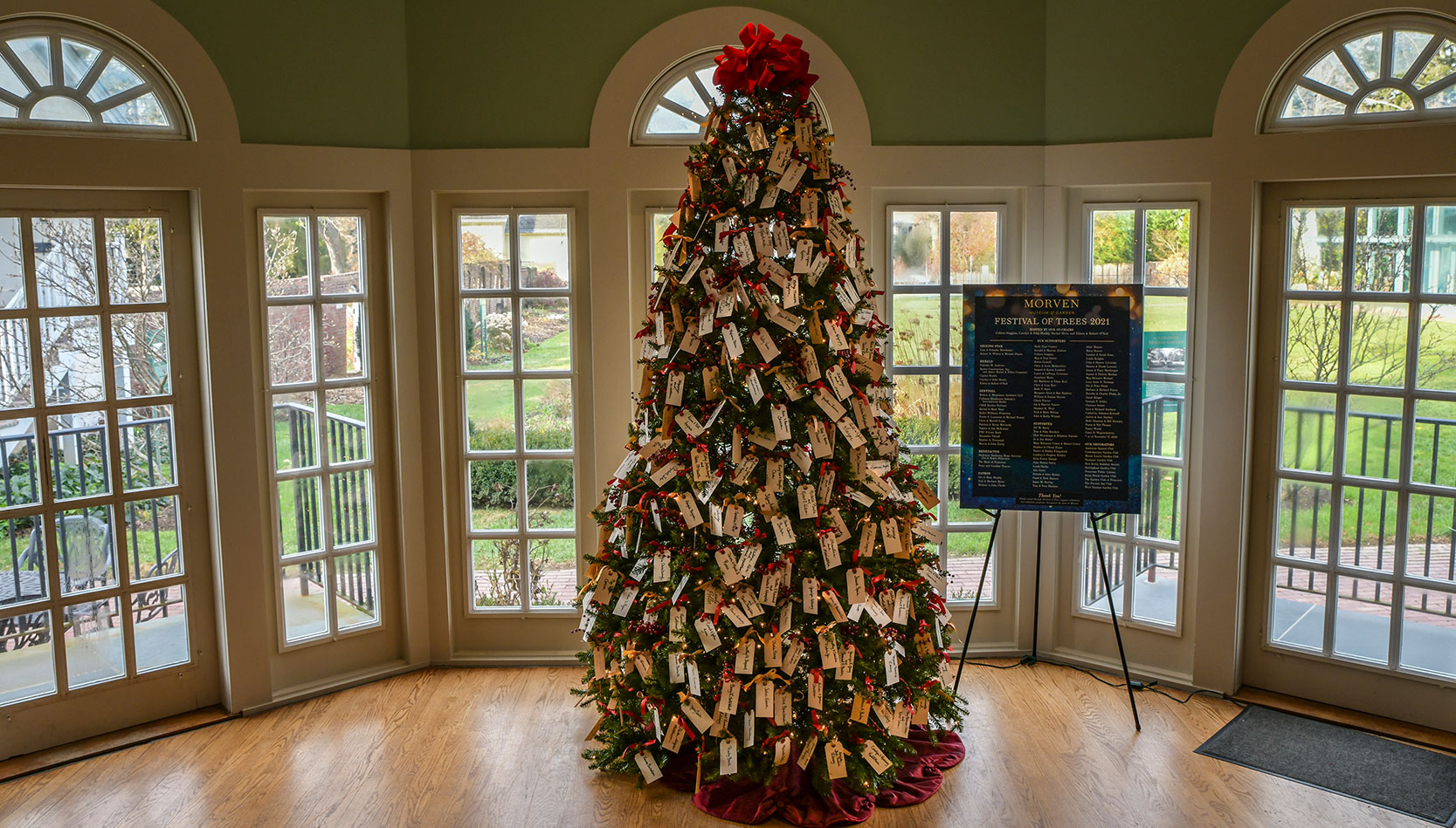 photo - A Christmas tree adorned with dozens of tags with peoples names stands in the middle of a green room. The rear garden area can be seen through windows and doors surrounding the display.