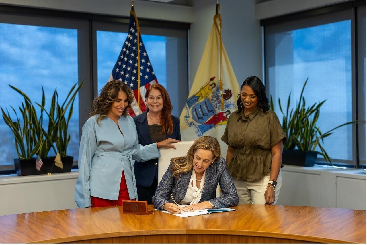 Governor Mikie Sherrill signing a bill at the desk with women and flags behind her