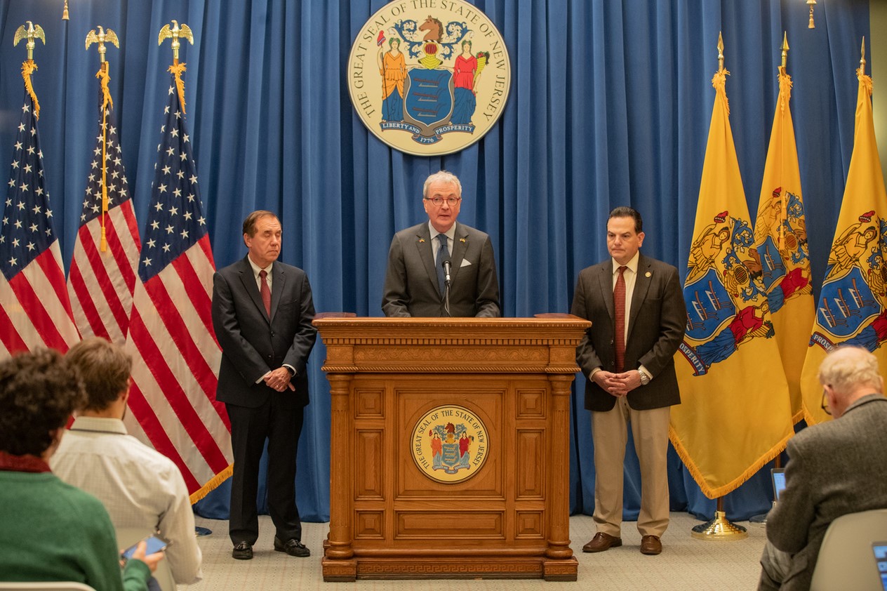 Photo: Governor Murphy is giving a speech with U.S. and NJ flags on the background and Speaker Coughlin and Senate President Scutari by his side