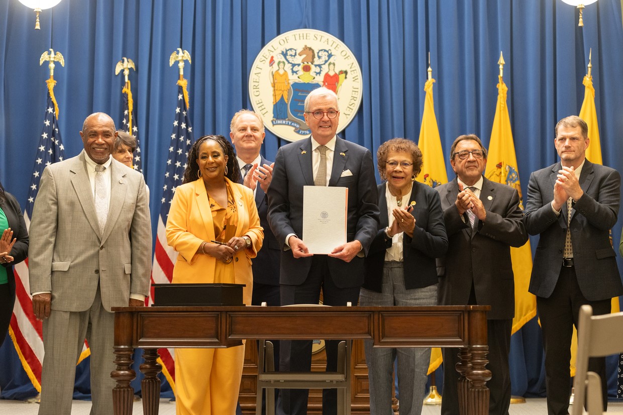 Photo: Governor Murphy is holding a signed Louisa Carman Medical Debt Relief Act with other people with NJ and America flags on the background