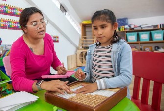 Female teacher of the blind and visually impaired and student approximately 8 years old  seated together in a classroom at a table working with Braille letter tiles