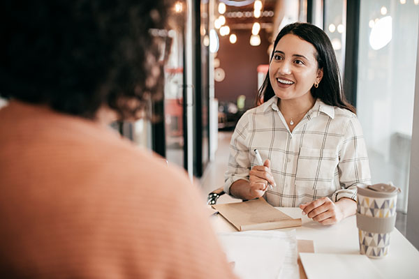 Woman with pen in hand smiling across table from another woman