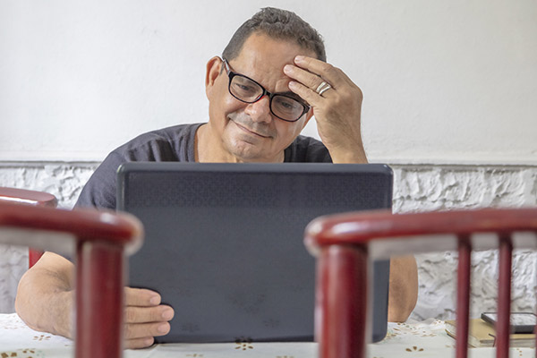 a senior man sitting at his kitchen table looking thoughtfully at a laptop computer