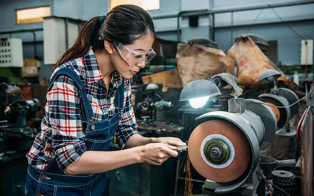 An Asian woman in a factory wearing safety goggles grinding a piece of metal