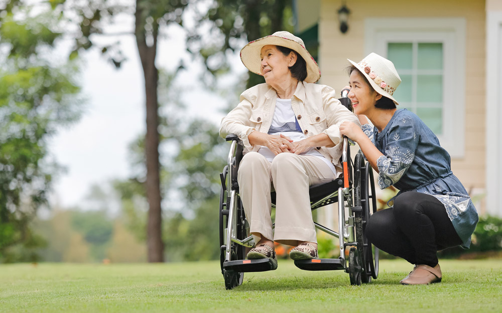 Smiling elderly Asian woman in a wheelchair in front of her house, with her daughter kneeling beside her 