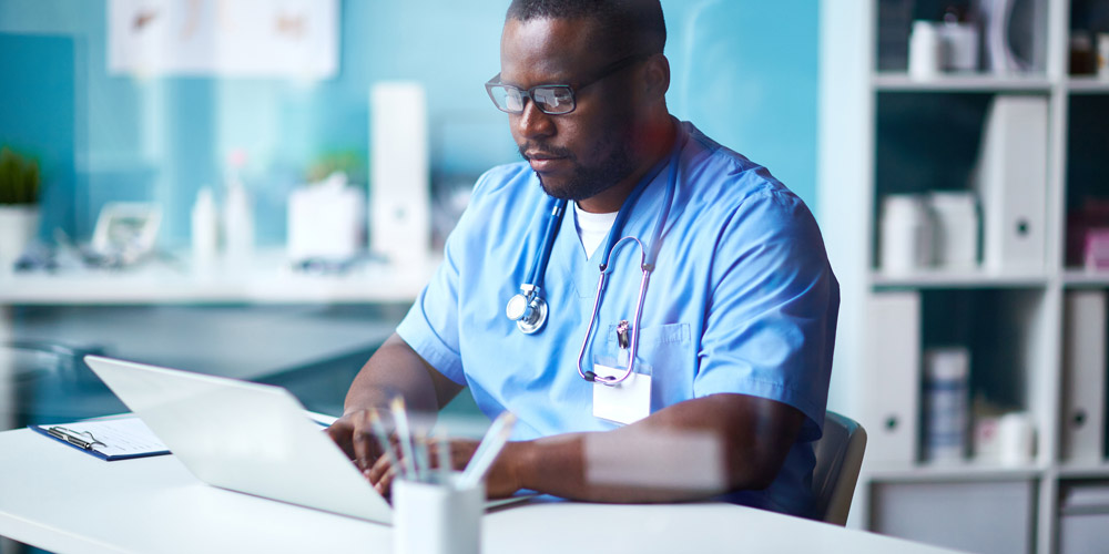 an African-American doctor sitting at his desk at his practice, typing on a computer