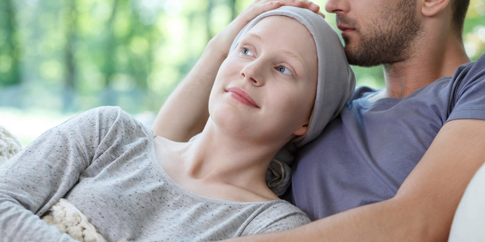 A woman who has lost her hair while undergoing chemotherapy smiling while leaning back against a man who is embracing her and holding the top of her head