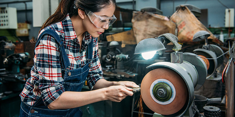 An Asian-American woman in a factory wearing safety goggles grinding a piece of metal