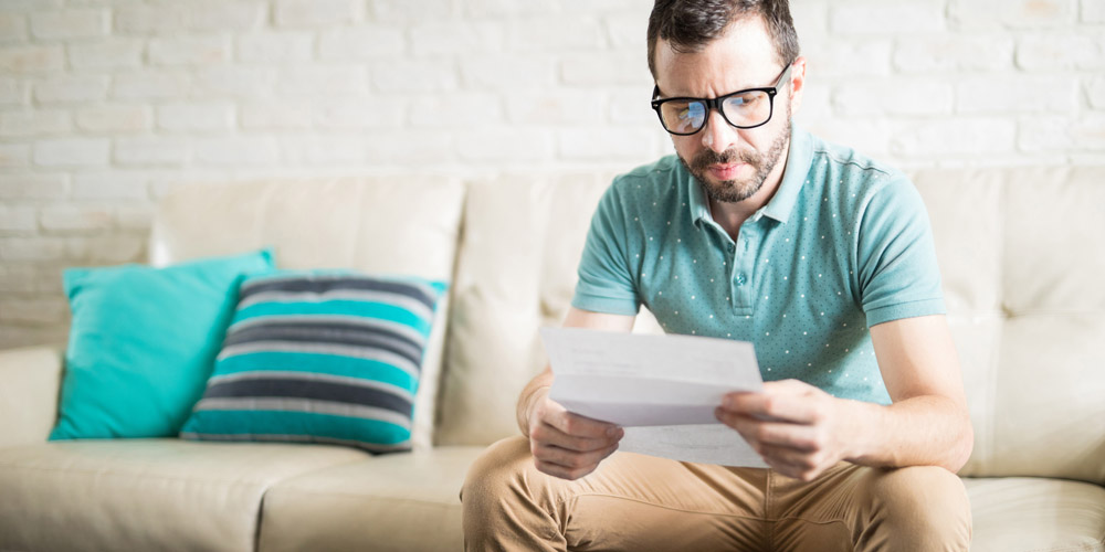 A man sitting on a sofa opening mail, looking confused