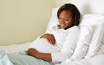 A pregnant African-American woman sitting on a bed, smiling and cradling her arms around her belly