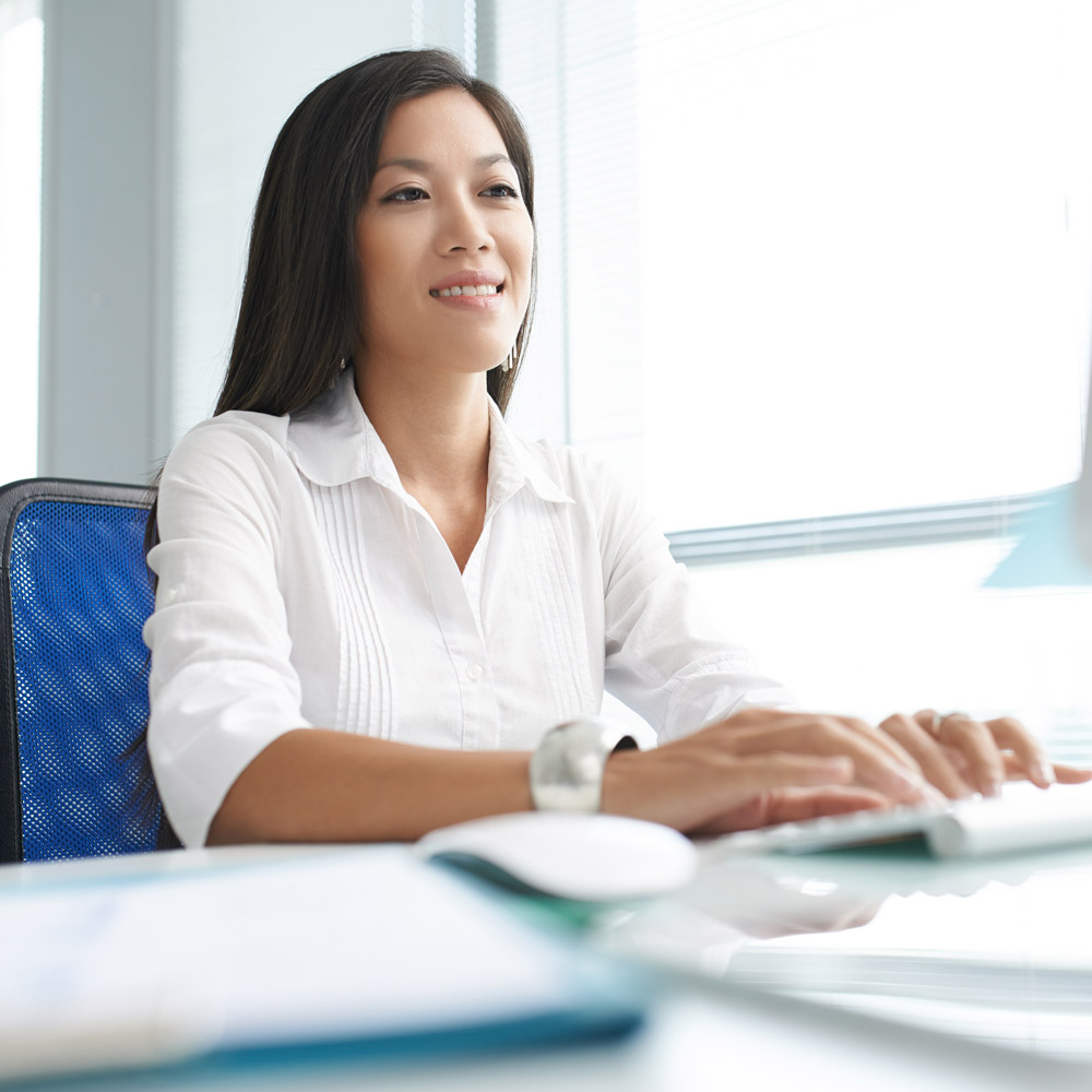 An Asian woman sitting at an office computer desk, smiling