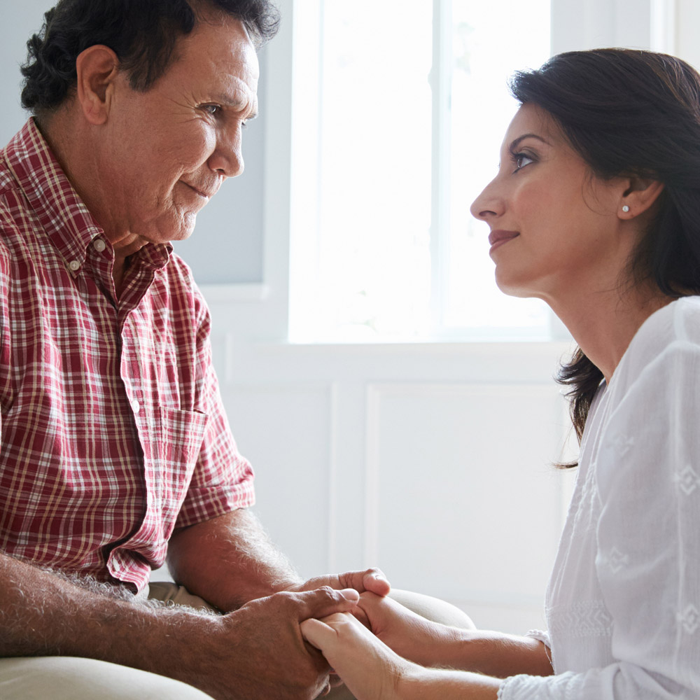 An elderly man sitting on the edge of a bed and smiling, with his daughter kneeling in front of him, holding his hands and smiling at him