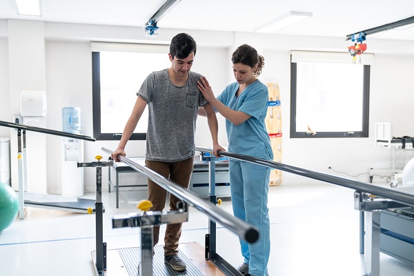 Focused male patient at physical therapy walking with the help of parallel bars and therapist next to him