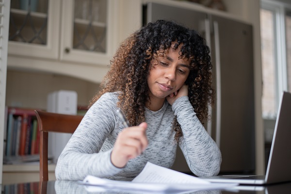 A young woman sits at her kitchen counter alone and fills out forms with a pen. Her laptop is open on the counter.