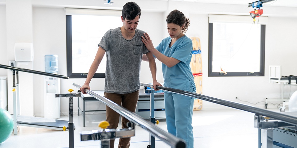 Focused male patient at physical therapy walking with the help of parallel bars and therapist next to him