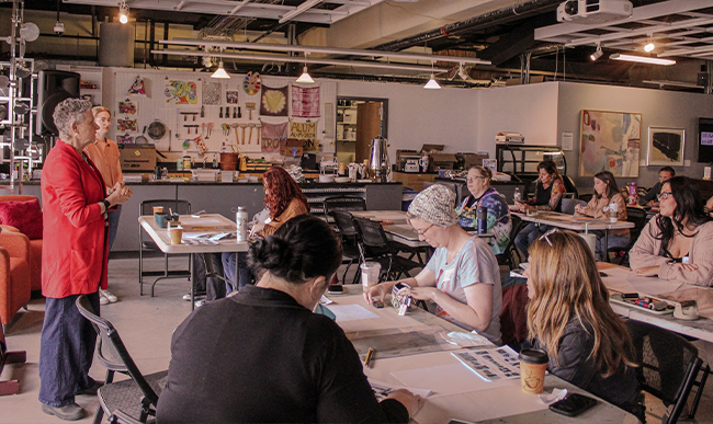 Group of people seated classroom-style in a creative studio space listening to a speaker at the front of the space.
