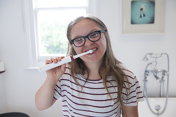 Woman with an intellectual disability standing in her bathroom brushing her teeth. She has long blonde hair and is wearing a white and burgundy striped shirt. She is wearing dark-framed glasses. She is holding a white toothbrush with a red strip. Her bathroom has white walls, and there is a white bathtub to her right with a picture frame above it. Behind her is a window.